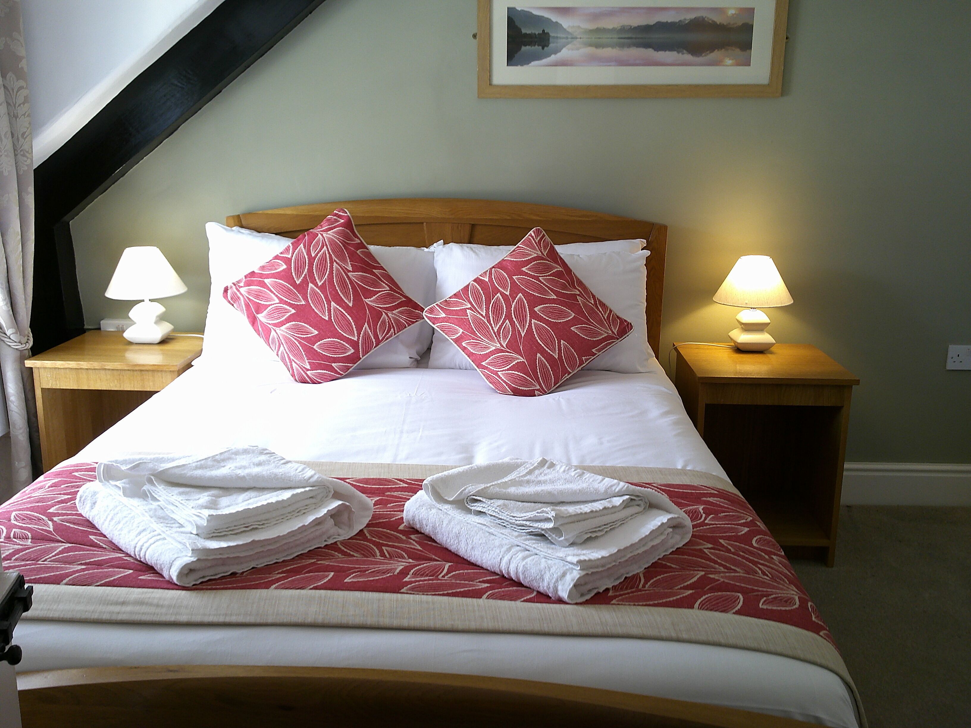 A neatly made bed with two pillows, two bedside lamps, and two folded towels on a bed with a red and white patterned bedspread.