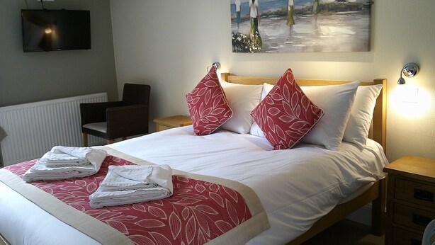 A neatly made bed with red and white linens, a painting of a beach scene above it, and a television mounted on the wall. - The Crown Hotel (Corwen)