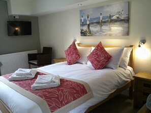 A neatly made bed with red and white linens, a painting of a beach scene above it, and a television mounted on the wall. - The Crown Hotel (Corwen)