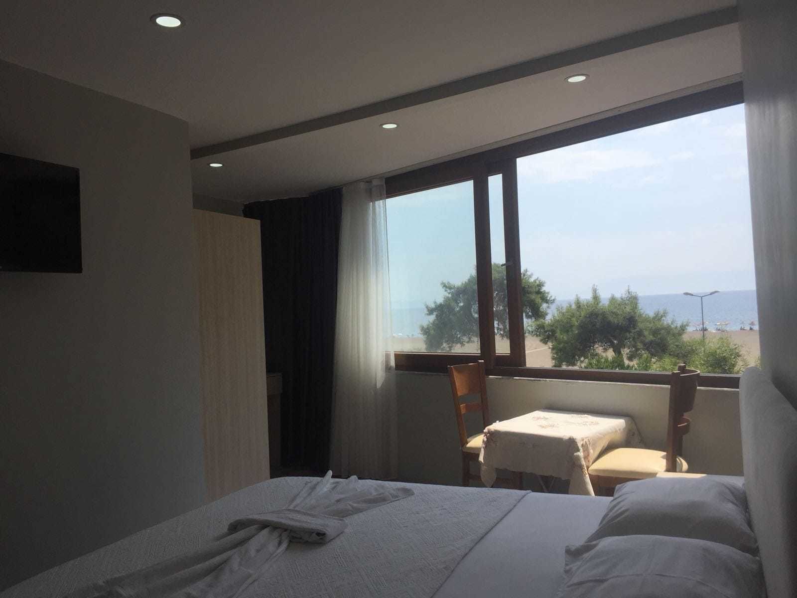 A hotel room with a bed, a small table, and a chair. There is a large window providing a view of the beach and the sea.