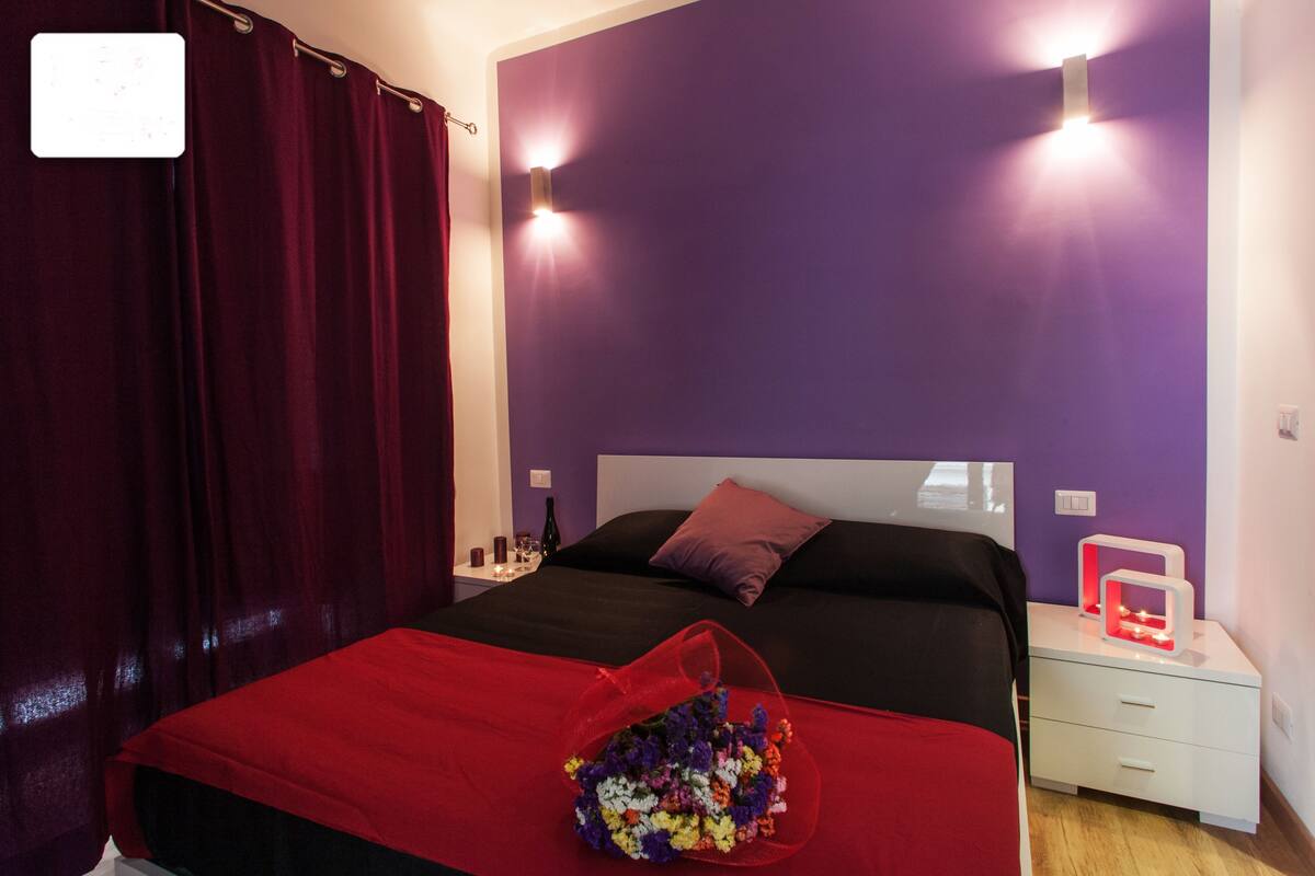 Ein Schlafzimmer mit roter Bettdecke, einem Blumenstrauß, einem Nachttisch mit Lampe und einer violetten Wand.