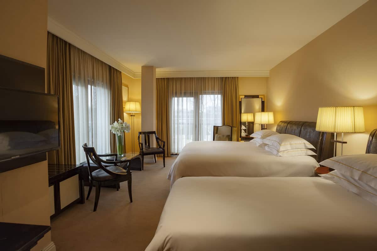 A hotel room with two beds, a desk with a chair, a TV, and lamps.