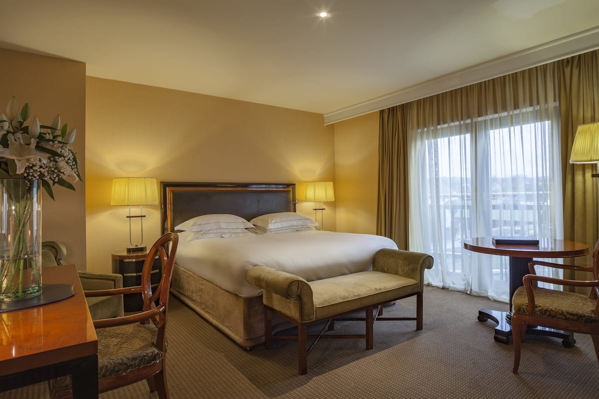 A hotel room with a large bed, two bedside tables with lamps, a wooden desk with a chair, and a view of the outdoors through the window.