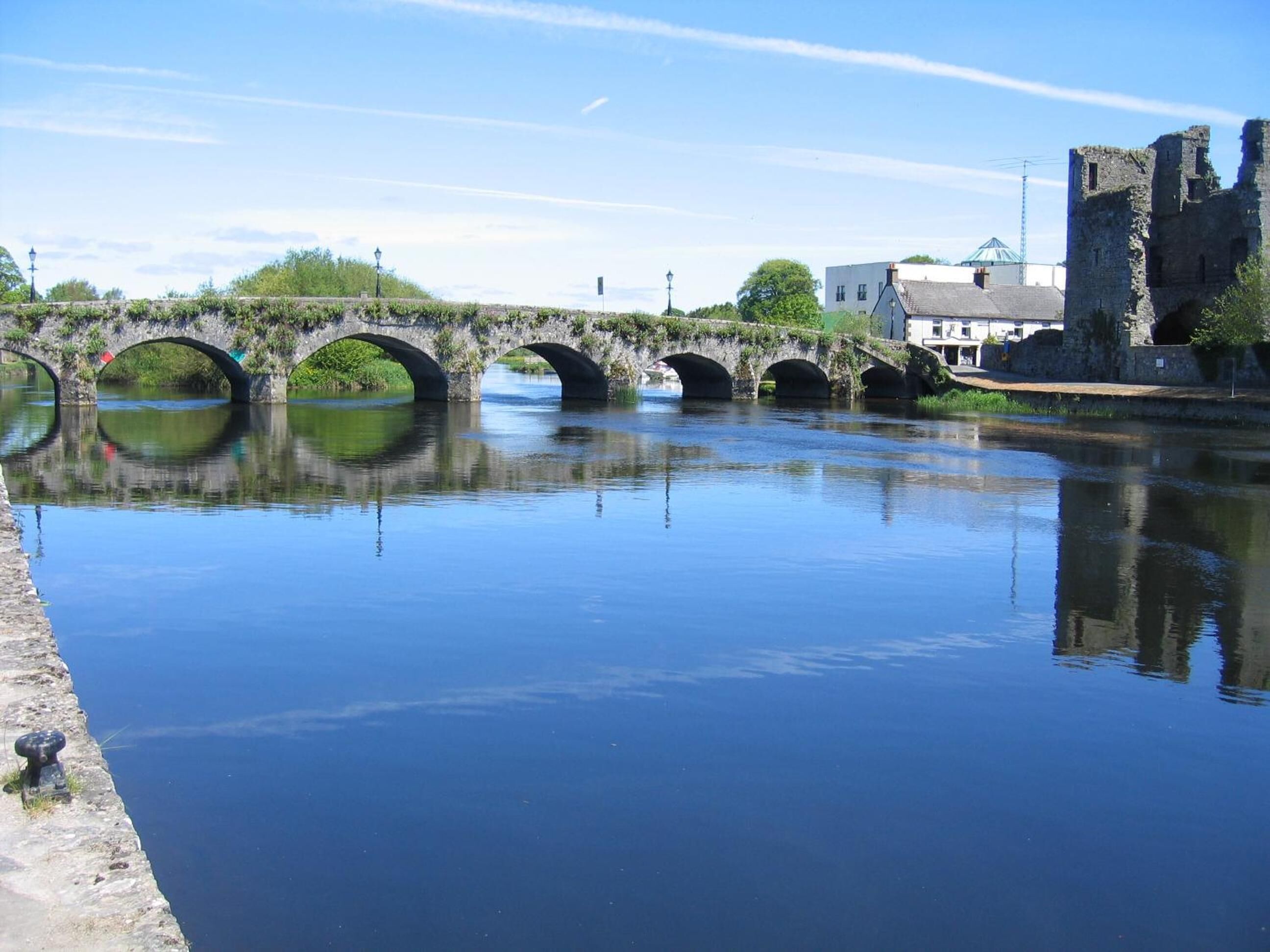A stone bridge over a calm river with a historic building in the background.