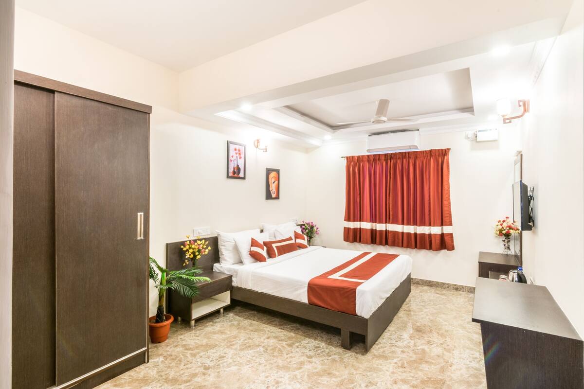 Une chambre d’hôtel avec un lit, un bureau, une armoire, une plante en pot et une fenêtre avec des rideaux.