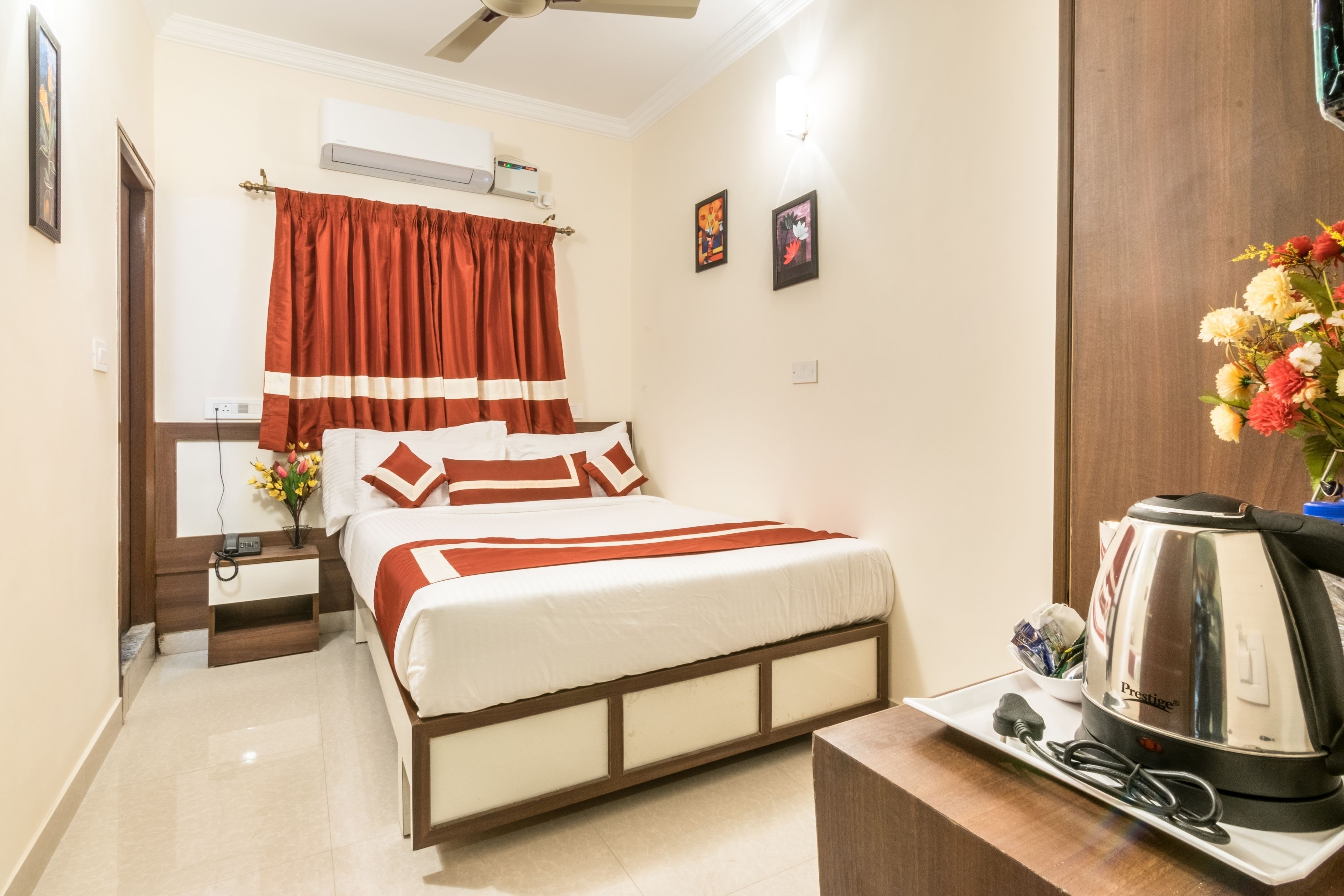 Une chambre d’hôtel bien rangée, avec un lit, une table de chevet sur laquelle se trouvent un réveil et un vase de fleurs, une machine à café et un climatiseur fixé au mur.