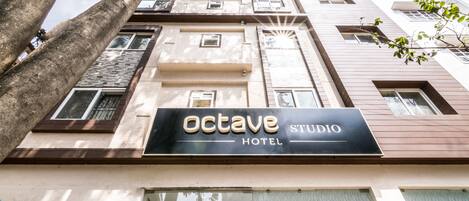 Exterior del Octave Studio Hotel, con un letrero llamativo, varios ventanales y un árbol en primer plano.