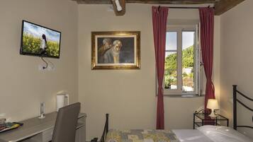 A bedroom with a bed, a desk, a chair, a television, a painting, and a window with curtains.