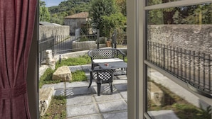 Una habitación con un ventanal, vista a un patio con mesa y sillas, y una pared de piedra.