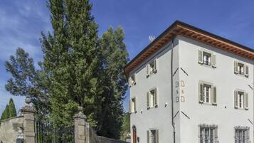 A two-story building with a white facade and a red-tiled roof. There are multiple windows with shutters, and a large tree is visible to the left.