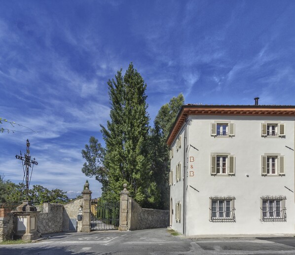Un edificio blanco con múltiples ventanas y contraventanas, un portón de piedra y un gran árbol en primer plano.