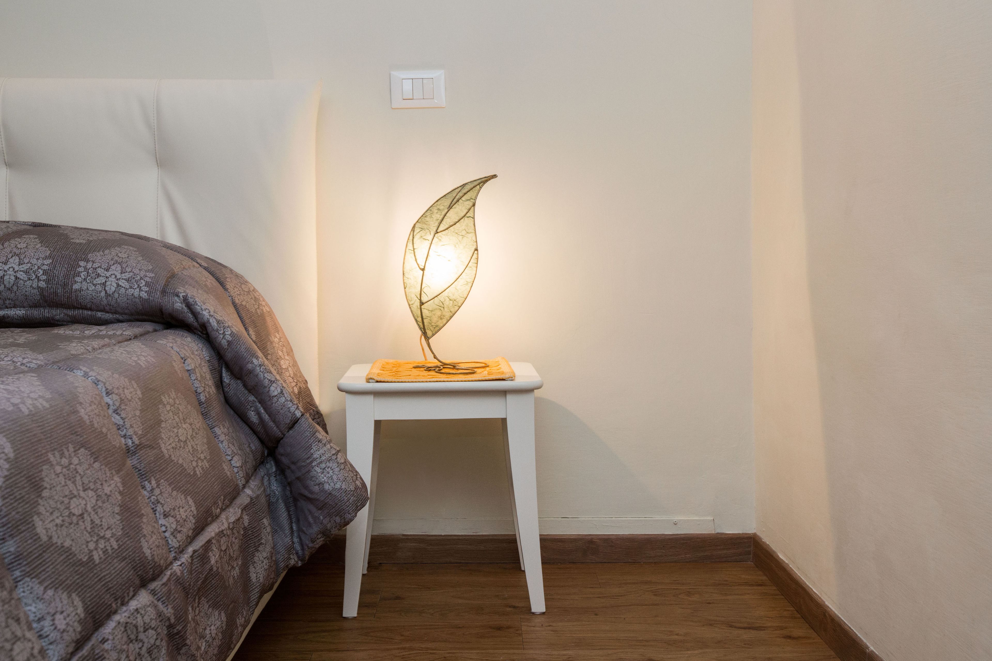 A bedroom with a bed, a small white nightstand with a leaf-shaped lamp, and a wooden floor.