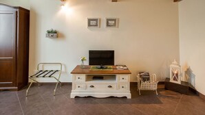 A room with a wooden ceiling, a white cabinet with drawers, a television, a small table with a vase, a wicker chair, and a wall-mounted shelf with a plant. - Charming Suite Cavour Heart of Florence (Florence)