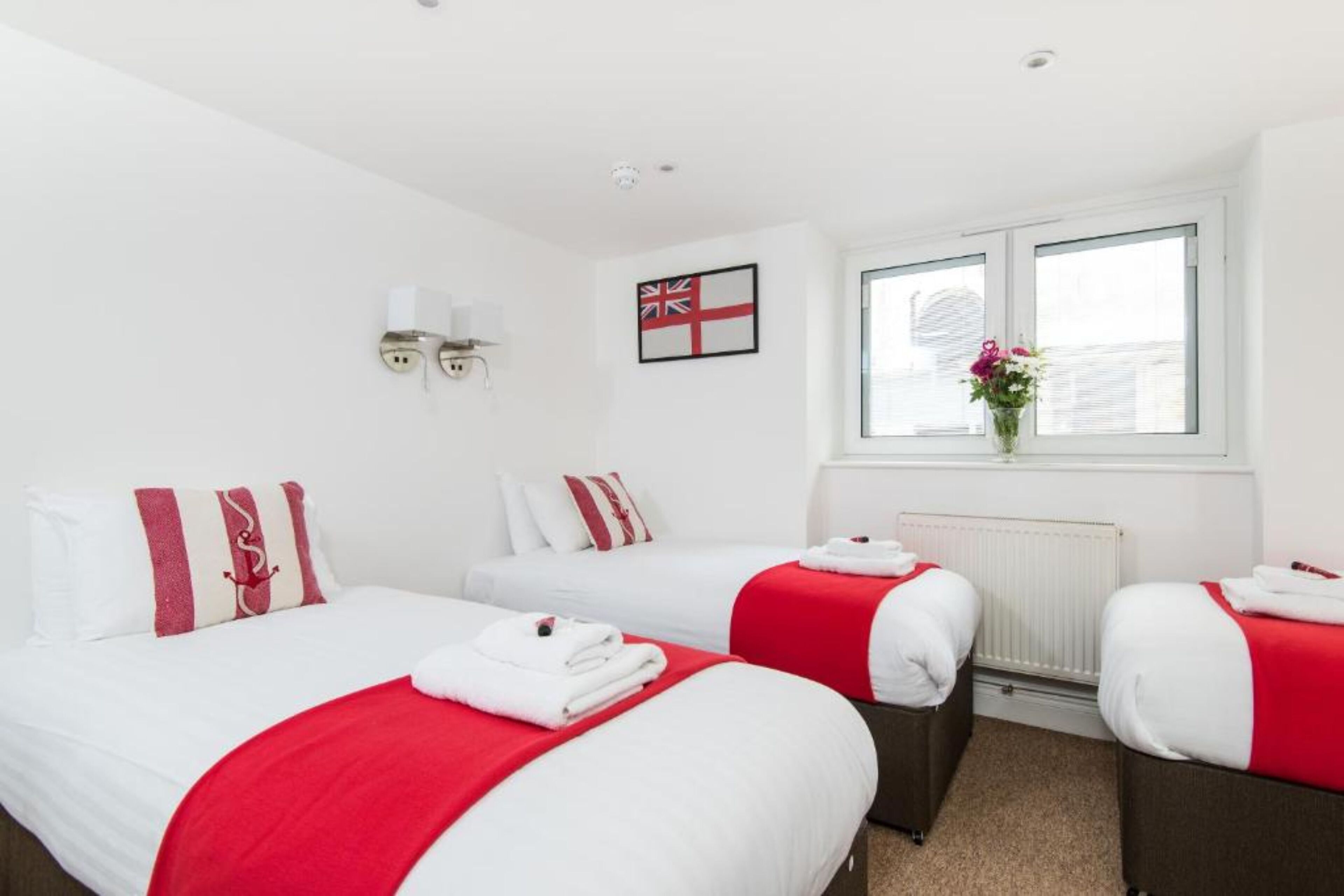 A hotel room with two beds, each with a red and white bedspread, white pillows, and a red and white striped pillow. There is a window with a view of a building, a radiator, and a wall-mounted flag.