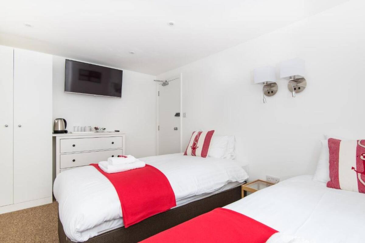 A hotel room with two beds, white walls, a flat-screen TV, a small table with a kettle, and a nightstand.