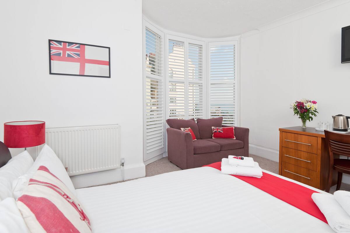 A hotel room with a bed, a red and white striped pillow, a brown sofa with red cushions, a wooden dresser, and a flag on the wall.