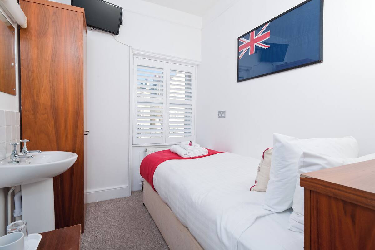 A hotel room with a wooden wardrobe, a white sink, a bed with white bedding, a red blanket, and a British flag on the wall.