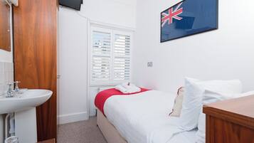 A hotel room with a wooden wardrobe, a white sink, a bed with white bedding, a red blanket, and a British flag on the wall.