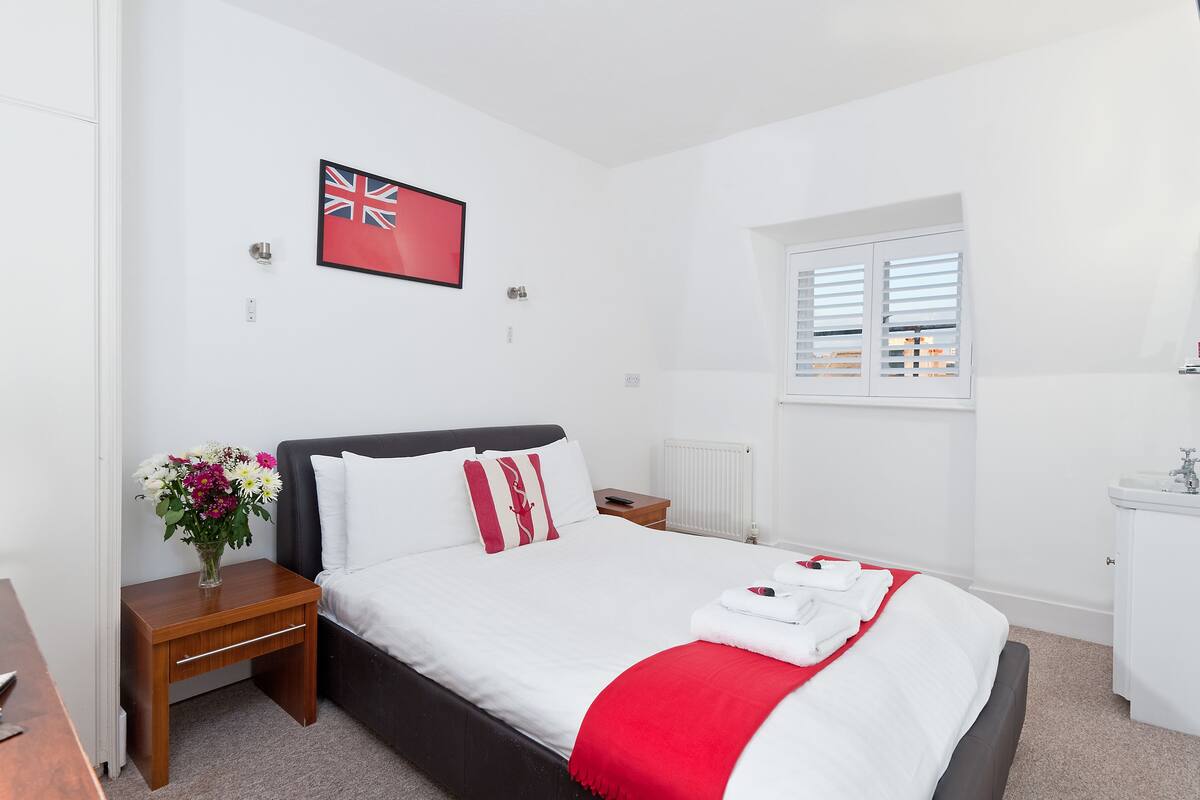 A hotel room with a bed, a nightstand with a vase of flowers, a window with blinds, and a small white cabinet.