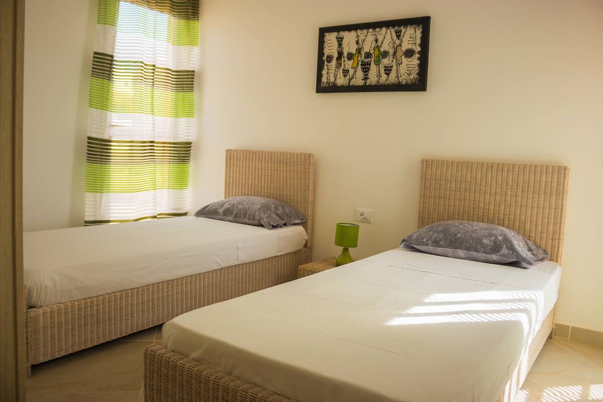 Two single beds with wicker headboards, a green nightstand, and a framed artwork on the wall.
