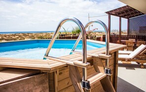 A wooden pool with metal ladders, lounge chairs, and a view of the beach. - Antonio Sousa Beach 2 Bedroom Apartment (Sal)