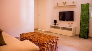 Una sala de estar con un sofá, un reposapiés de mimbre, un televisor de pantalla plana sobre un soporte blanco, una cortina verde estampada y una puerta blanca.
