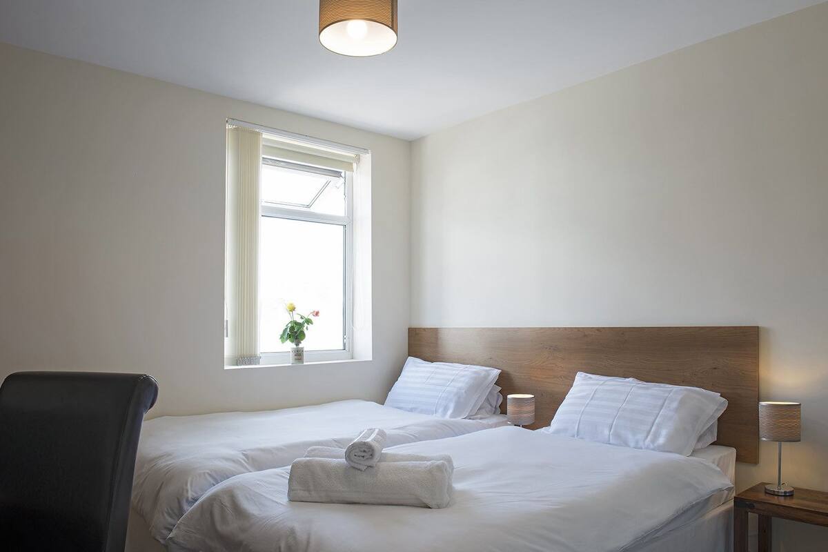 A hotel room with two beds, a window with a view, a chair, and a bedside table with a lamp.