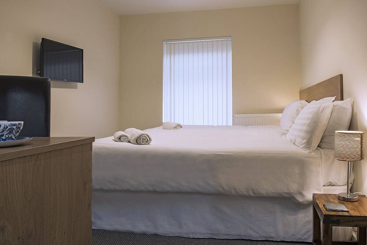 A hotel room with a bed, a bedside table with a lamp, a flat-screen TV mounted on the wall, and a window with blinds.