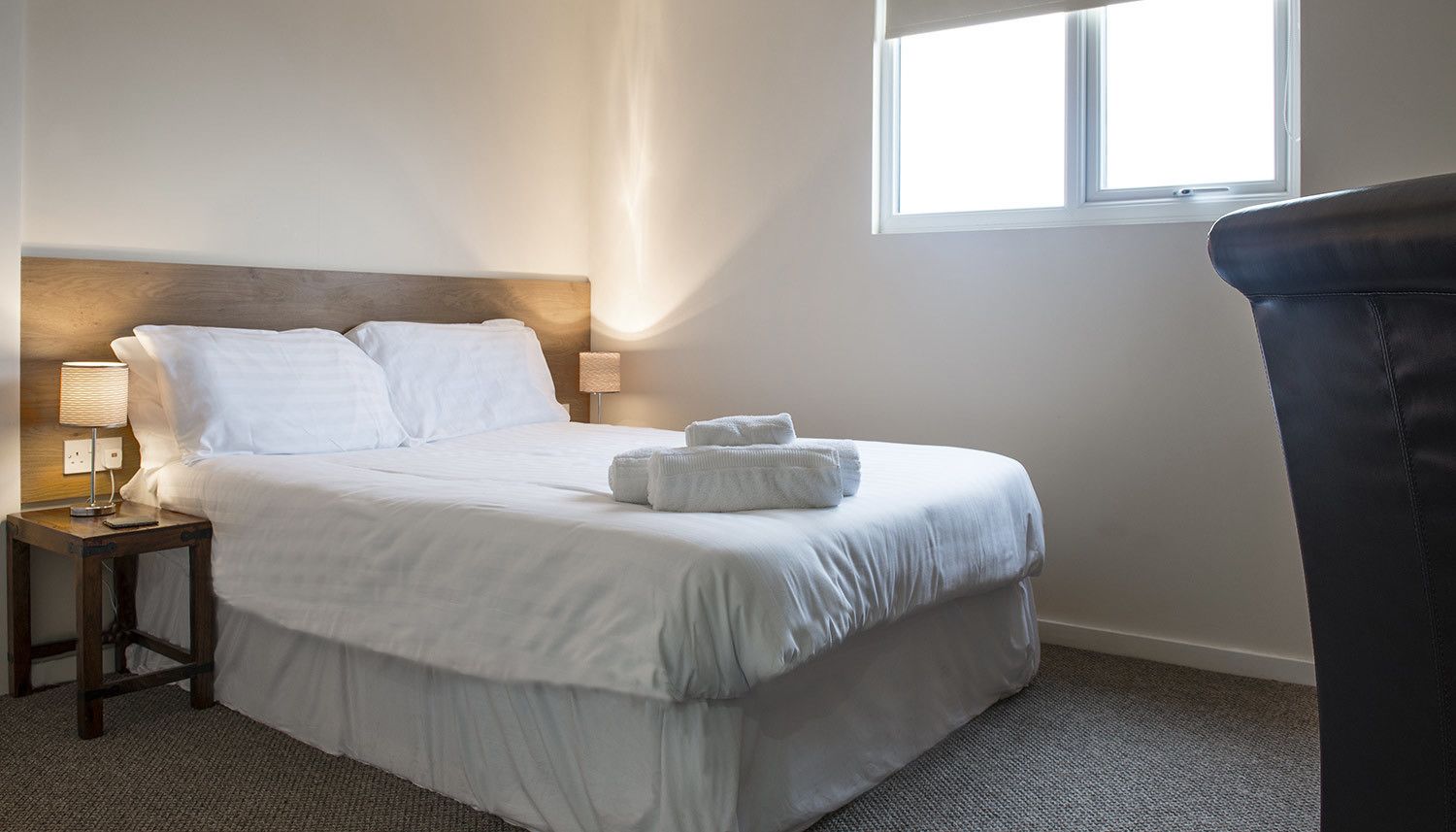 A neatly made bed with white linens and two pillows, a bedside lamp, and a window with daylight.