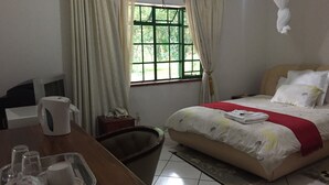 A hotel room with a bed, a desk with a telephone and a tray with glasses and a cup, a window with curtains, and an air conditioning unit. - Divine Guest House (Livingstone)