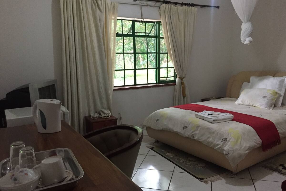 A hotel room with a bed, a desk with a telephone and a tray with glasses and a cup, a window with curtains, and an air conditioning unit.