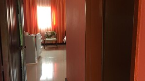 A hallway leading to a room with a chair and a table, a refrigerator, and orange curtains. - Divine Guest House (Livingstone)
