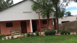 A two-story building with a red and white facade, a porch with a bench, and a small garden area. - Divine Guest House (Livingstone)