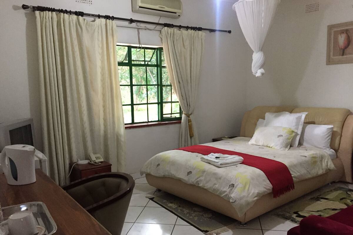 A hotel room with a bed, a bedside table, a chair, a window with curtains, a bedside lamp, and a wall-mounted air conditioner.
