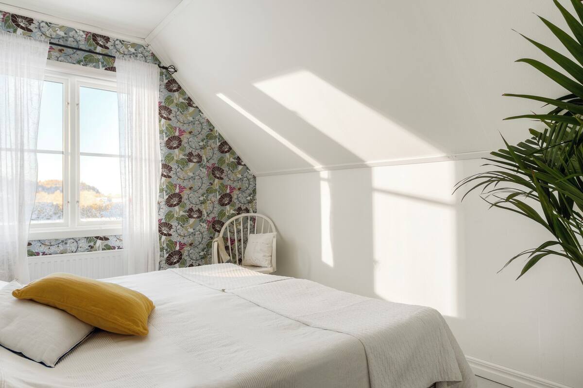 A bedroom with a bed, a chair, a window with floral curtains, and a plant.