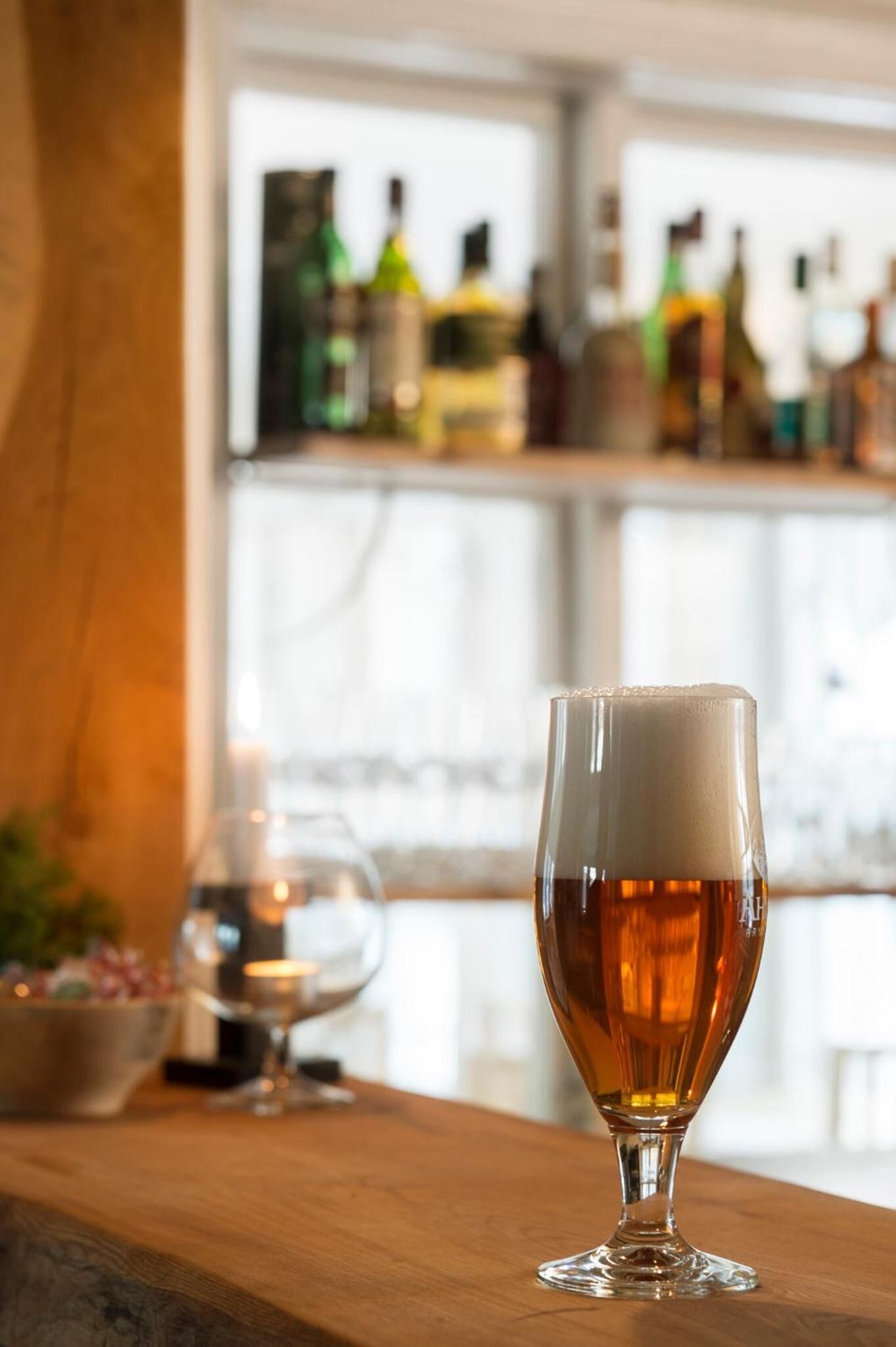 A glass of amber beer on a wooden bar counter with a blurred background of bottles and a window.