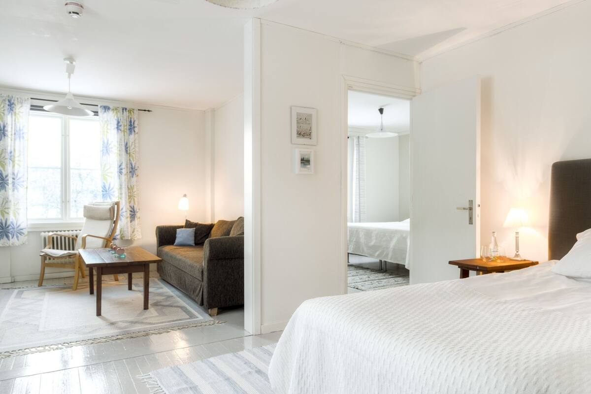 A hotel room with a bed, a sofa, a table, and a chair. There is a window with curtains, a lamp, and a picture on the wall.