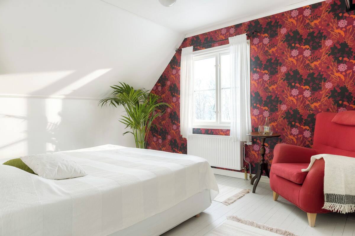 A bedroom with a bed, a red armchair, a potted plant, and a window with curtains.