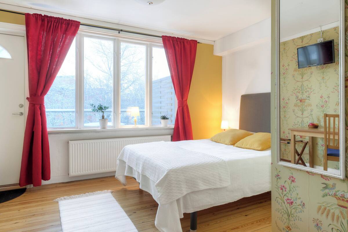 A bedroom with a bed, a window with curtains, a radiator, a wooden floor, and a television on the wall.