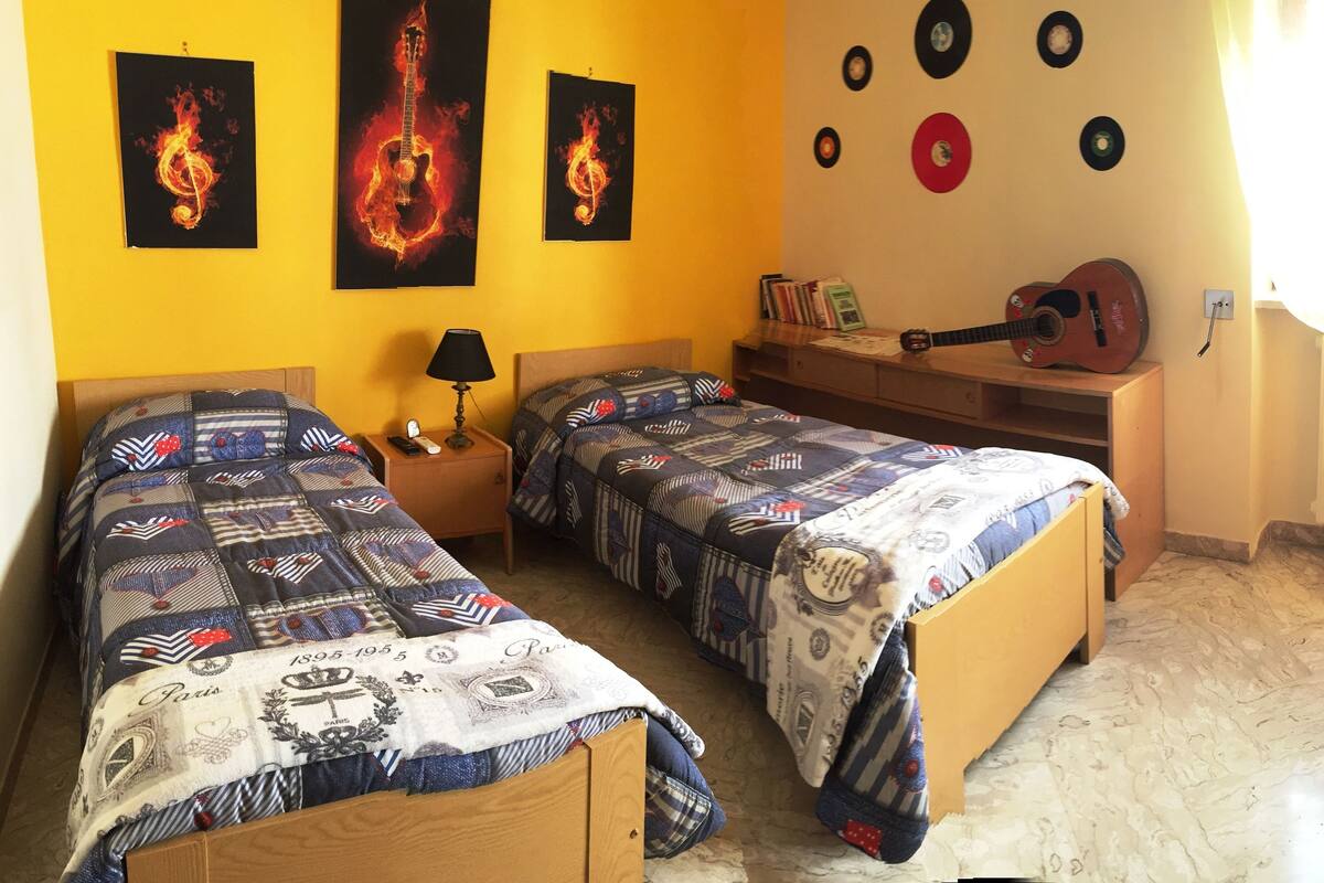 A room with two beds, a nightstand, a guitar, and wall decorations including records and framed artwork.