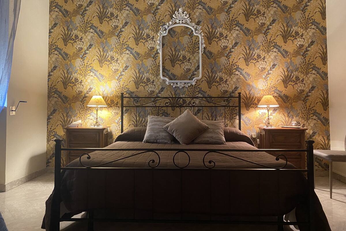 A bedroom with a bed, two bedside lamps, a mirror, and floral wallpaper.
