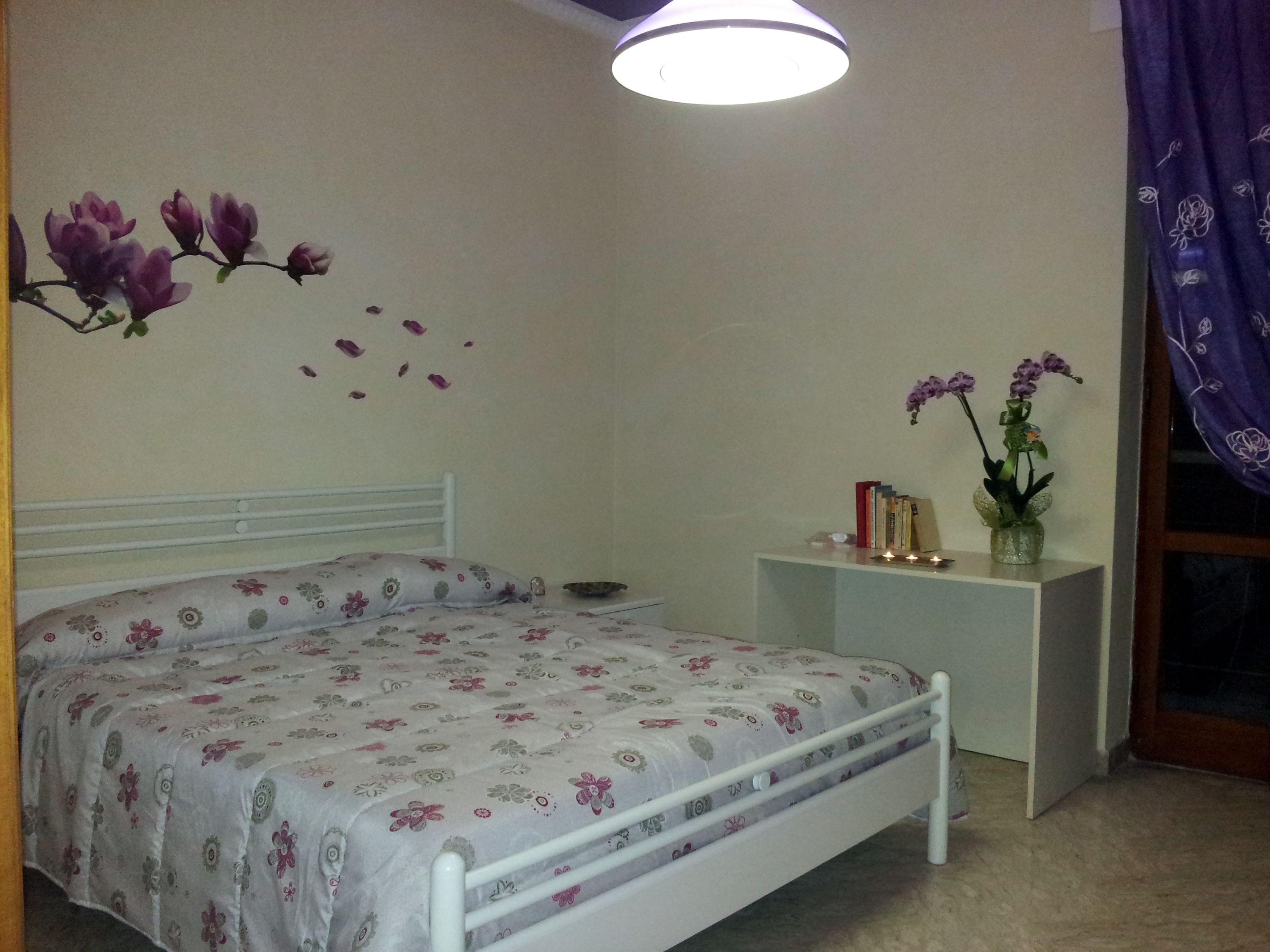 Un dormitorio con cama, mesita de noche, un jarrón con flores y un adorno en la pared.