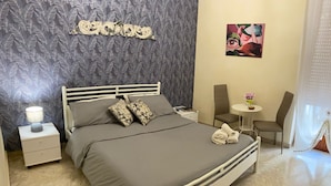 A neatly made bed with a grey bedspread and two patterned pillows, a bedside table with a lamp, a small round table with a vase, and a chair. - La Dimora di Vesta (Lecce)