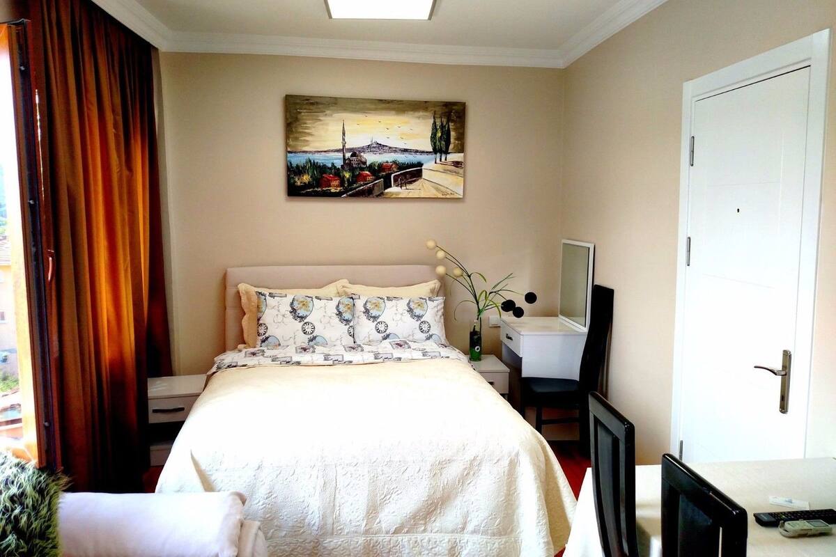 A hotel room with a bed, a bedside table with a mirror, a desk with a computer, and a painting on the wall.