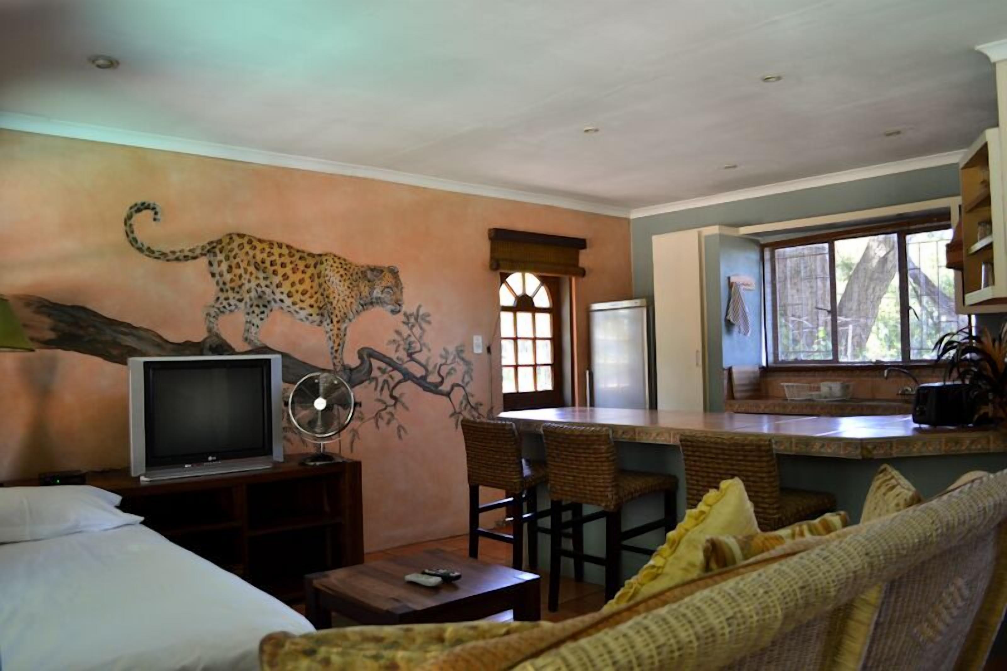 A room with a bed, a television, a dining area with chairs, a kitchen area with a refrigerator, and a leopard mural on the wall.