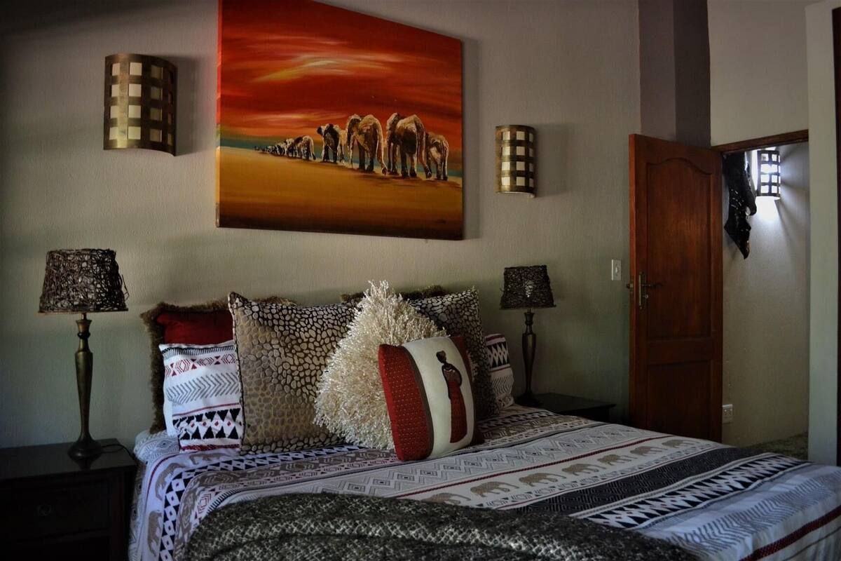 Ein Schlafzimmer mit einem großen Bett, gemusterten Betttextilien, zwei Wandlampen, einem Elefantenbild und einer Holztür.