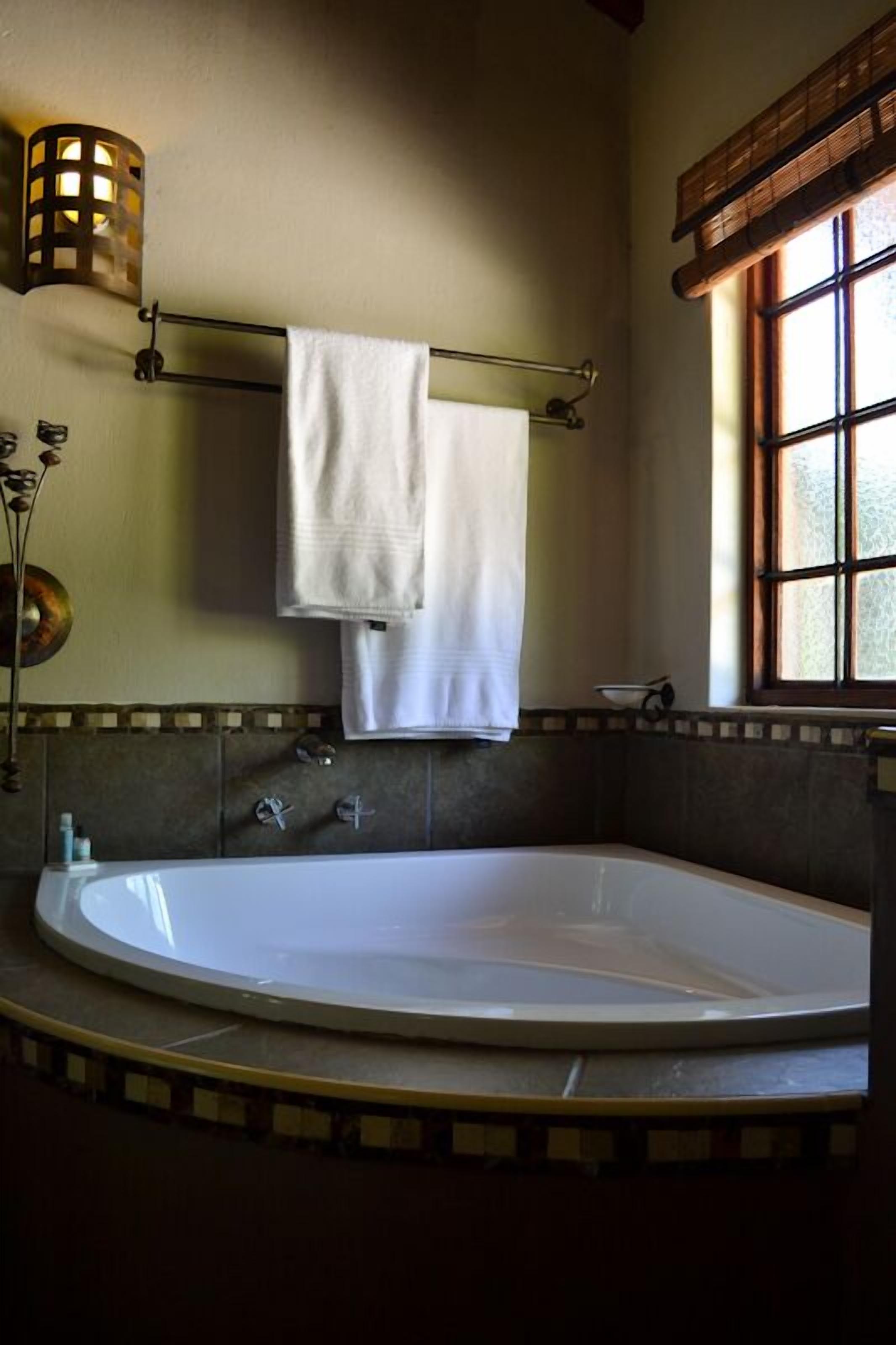 A bathroom with a large bathtub, a towel rack with two white towels, a wall-mounted light fixture, and a window with a view of greenery.