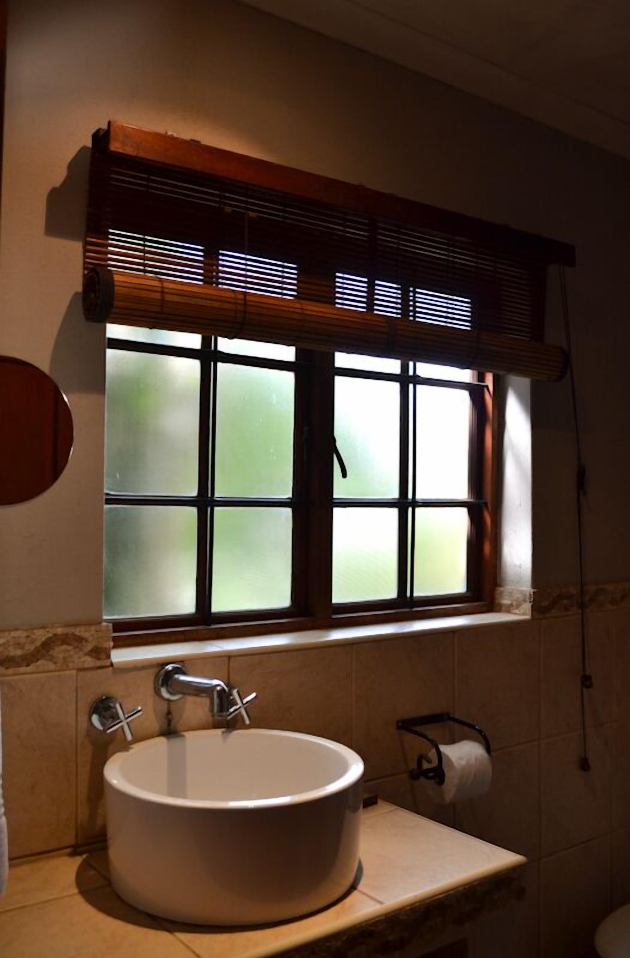 A bathroom with a round sink, a window with blinds, and a toilet paper holder.