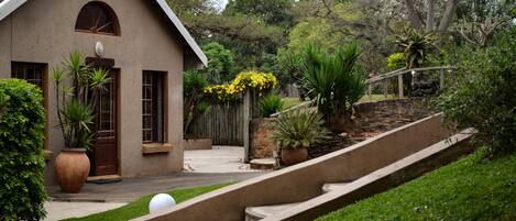 Una casa con techo inclinado, puerta principal y un pequeño porche. Un camino pavimentado conduce a la casa, y un área verde con algunas plantas en macetas.