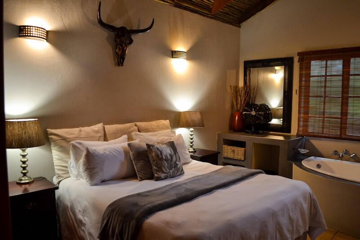 Ein Schlafzimmer mit einem Bett, Nachttischlampen, einem Wandspiegel, einem großen Spiegel, einem Fenster mit Jalousien und einem an der Wand befestigten Bauschädel.